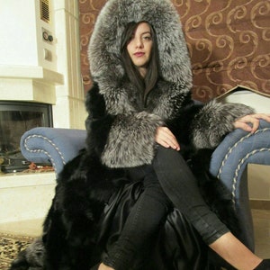 Puede incluir: Una mujer lleva un abrigo largo de piel negra con capucha de piel de zorro plateado. Está sentada en un sillón azul con las piernas cruzadas. Lleva pantalones vaqueros negros y tacones negros.