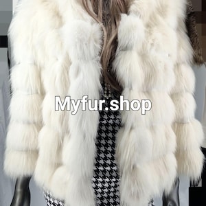 May include: A white fur jacket with a long, fluffy collar. The jacket is made of fox fur and has a layered design. The jacket is being worn by a mannequin with a black and white houndstooth skirt. The text "Myfur.shop" is visible on the jacket.