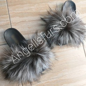 May include: A pair of black faux fur slides with a fluffy silver fox fur pom pom on each slide. The slides are on a light brown tiled surface. The text "Angellsfurs.com" is visible in the image.