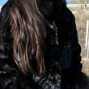 May include: A black fur jacket with a fur collar. The jacket is worn by a person with long dark hair and round sunglasses.