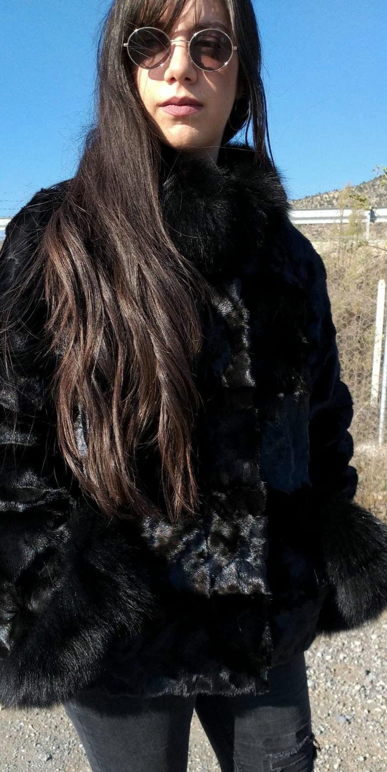 May include: A black fur jacket with a fur collar. The jacket is worn by a person with long dark hair and round sunglasses.
