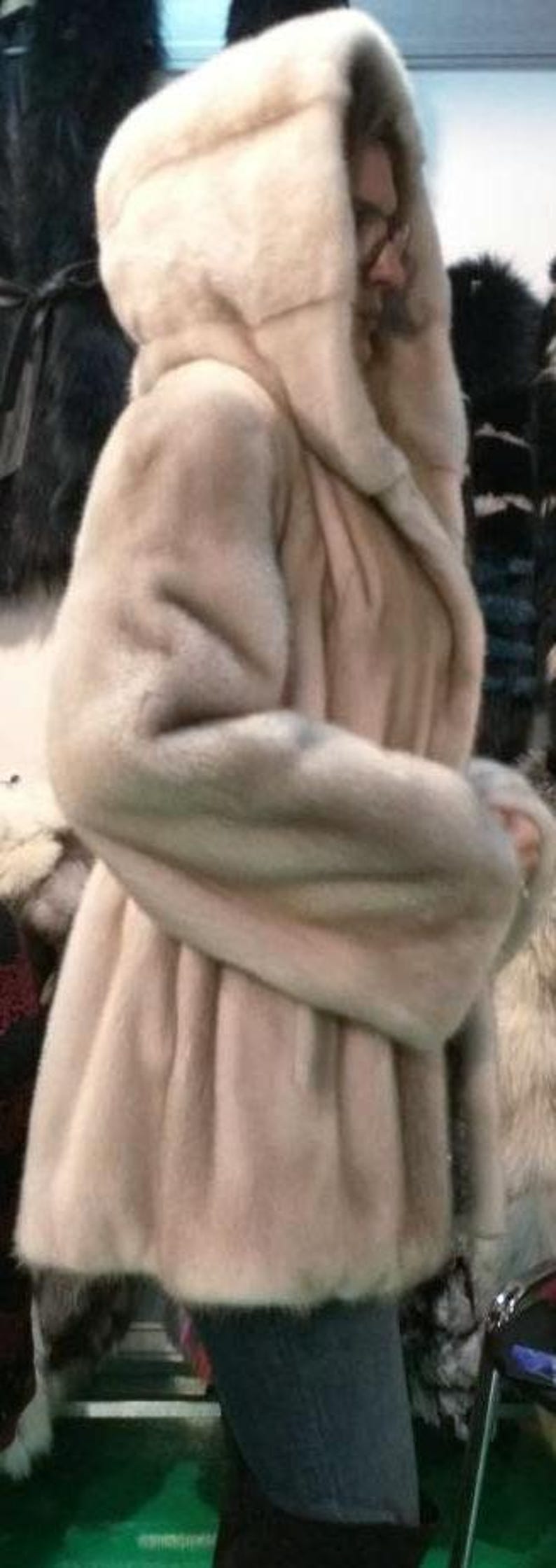 May include: A light beige fur coat with a large hood. The coat is made of soft, fluffy fur and has a long, flowing style.