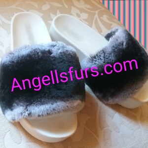 May include: A pair of white faux fur slides with black fur trim. The slides have a white sole and the fur is soft and fluffy. The text "Angellsfurs.com" is printed on the fur.