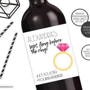 May include: A white wine bottle label with black text that reads "ALEXANDRA'S last fling before the ring!" with a pink diamond ring illustration. The label also includes the date "JULY 20.2019" and the hashtag "#YOURHASHTAGHERE".