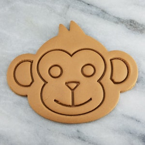 May include: A brown cookie cutter shaped like a monkey face. The cutter has a simple design with two large eyes, a small nose, and a smiling mouth.