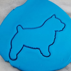 May include: A blue cookie cutter in the shape of a Norwich Terrier dog. The cutter is pressed into blue fondant.