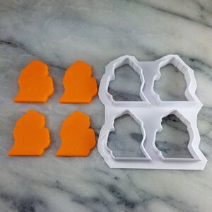 May include: Four orange cookie cutters in the shape of a house, next to a white plastic cookie cutter with four house-shaped molds.