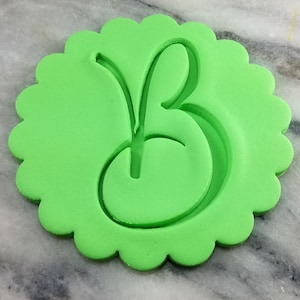 Könnte beinhalten: Grüner Plastik-Ausstecher in Form eines gewellten Kreises mit einem ausgeschnittenen kursiv geschriebenen Buchstaben "B" in der Mitte.