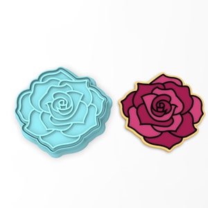 May include: A blue plastic cookie cutter shaped like a rose, next to a pink and red cookie in the shape of a rose.