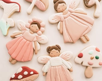 Op de afbeelding: Een set van negen gedecoreerde suikerkoekjes met feeën- en paddenstoelmotieven. De koekjes zijn versierd met roze, wit, bruin en rood glazuur.