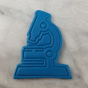 May include: A blue plastic cookie cutter shaped like a microscope. The microscope has a round base and a long, curved neck with a lens at the top.