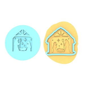 Op de afbeelding: Een blauwe plastic koekjesvormer in de vorm van een kerststal met een ster boven de stal. De vormer wordt getoond op een gele achtergrond.