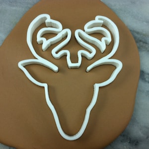 May include: White plastic cookie cutter in the shape of a deer head with antlers. The cutter is on a brown surface.