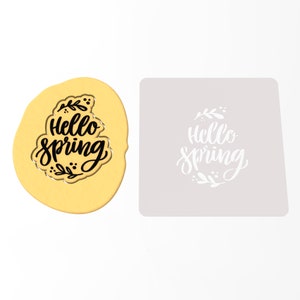 Op de afbeelding: Een gouden koekjesvormpje met de woorden "Hello Spring" in zwarte schrijfletters en een bloemmotief. Naast het koekjesvormpje is een witte sjabloon met hetzelfde ontwerp te zien.