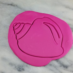 May include: A pink polymer clay imprint of a seashell cookie cutter. The seashell has a detailed spiral design.