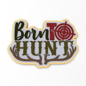 May include: A cookie decorated with the words "Born to Hunt" in green and black, with a red target symbol and brown antlers.
