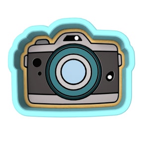 May include: A cartoon-style camera cookie cutter. The camera is gray with a teal lens and a light blue center. The cutter has a light blue outline with a gold-colored interior.