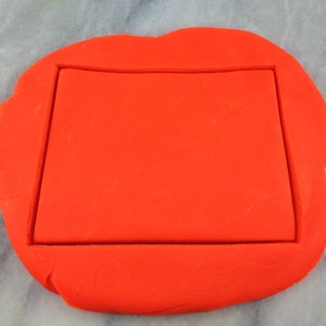 Puede incluir: Una masa de galleta de fondant naranja brillante con un recorte rectangular en el centro.