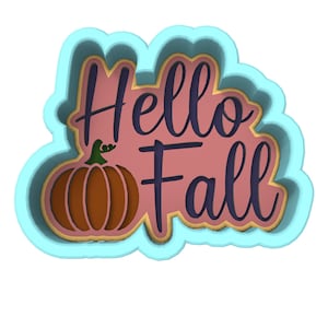 Op de afbeelding: Een lichtblauwe koekjesvormer met de woorden "Hello Fall" in paars en een bruine pompoen in het midden.