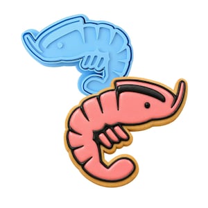 Puede incluir: Un cortador de galletas de plástico azul claro con forma de camarón colocado sobre una galleta con forma de camarón decorada con glaseado rosa y contornos negros. La galleta tiene un cuerpo curvado, una cabeza definida y patas pequeñas.