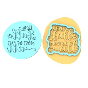 Op de afbeelding: Twee koekjesvormen, een blauwe en een gele, met de tekst "Ik hou van de herfst het meest" in een decoratief lettertype. De koekjesvormen zijn ontworpen om koekjes uit te steken in de vorm van de tekst.