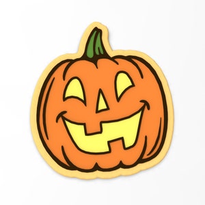 Puede incluir: Una galleta de calabaza naranja sonriente con una cara amarilla y un contorno negro. La galleta está decorada con glaseado amarillo para los ojos, la nariz y la boca.