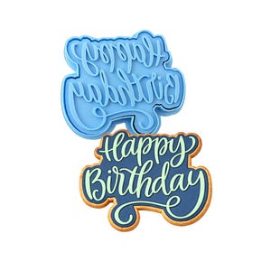 May include: A light blue plastic cookie cutter shaped like the words Happy Birthday in a cursive font, positioned above a matching cookie decorated with dark blue icing and light green lettering that reads Happy Birthday.