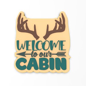 May include: A wooden sign with a beige background and a brown deer antler design at the top. The sign says "Welcome to our Cabin" in teal blue lettering with a black arrow pointing to the right.