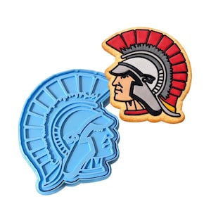 May include: A light blue cookie cutter and a decorated cookie in the shape of a Roman soldier's helmet. The cookie has red, white, black, and yellow details. The cookie cutter is made of blue plastic.