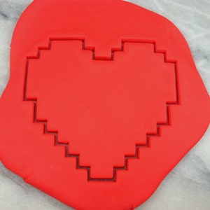 Könnte beinhalten: Ein leuchtend roter, pixeliger herzförmiger Ausstecher. Der Ausstecher besteht aus einem flexiblen Material und ist in einen roten Teig gedrückt. Das Design ist eine moderne Interpretation einer klassischen Herzform, zum Backen.