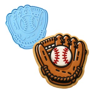 May include: A light blue plastic baseball glove cookie cutter alongside a decorated sugar cookie shaped like a baseball glove with a white baseball featuring red stitching, brown leather tones, and black piping details on the glove edges.