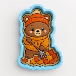 May include: A light blue cookie cutter featuring a brown bear wearing an orange knit hat and a matching orange sweater with a tree pattern. The bear is holding a metal rake and standing over a pile of orange, yellow, and red autumn leaves.