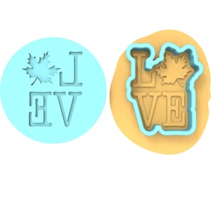 Op de afbeelding: Een blauwe plastic koekjesvormer met het woord "LOVE" en een esdoornbladontwerp. De vormer wordt naast een bruin deeg oppervlak getoond.