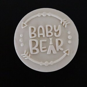 May include: White round cake topper with the text "BABY BEAR" in a whimsical font, surrounded by a decorative border of arrows and dots.