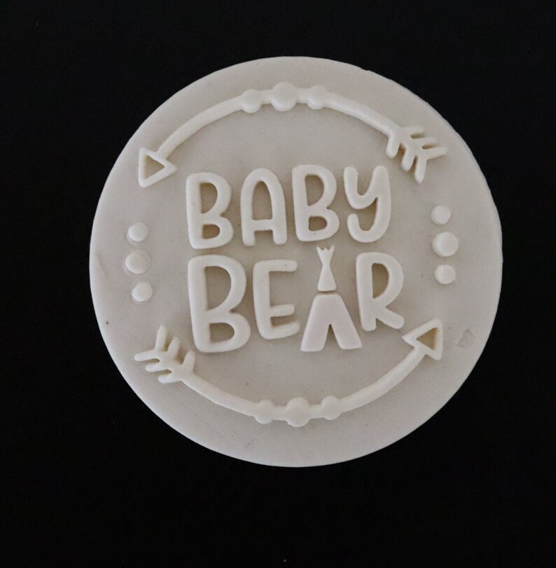 May include: White round cake topper with the text "BABY BEAR" in a whimsical font, surrounded by a decorative border of arrows and dots.