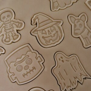 Puede incluir: Primer plano de una hoja de masa para galletas marr&oacute;n con varios cortadores de galletas con tem&aacute;tica de Halloween presionados en la masa. Los cortadores incluyen un fantasma, una calabaza, un esqueleto, un caramelo, un Frankenstein y un murci&eacute;lago.