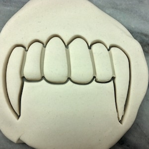 May include: A white plastic cookie cutter shaped like a set of vampire fangs. The cutter has five teeth and two large fangs.