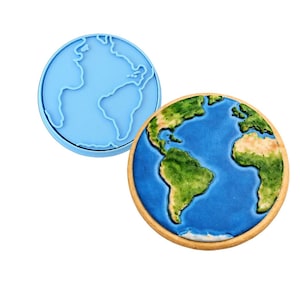 May include: A blue silicone cookie cutter and a decorated Earth-shaped cookie. The cookie has a blue ocean, green continents, and a brown border. The cookie cutter is a light blue outline of the Earth.