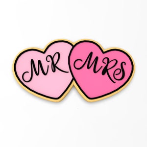 May include: Two pink heart-shaped cookies with gold outlines. The left cookie says "MR" and the right cookie says "MRS".
