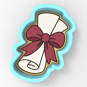 May include: A cookie cutter in the shape of a rolled-up scroll tied with a burgundy bow. The scroll is off-white, outlined in gold, and set within a light blue border. The design is a cartoon-style illustration.