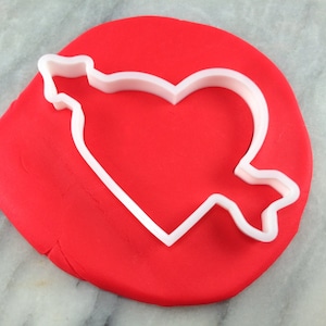 Puede incluir: Un cortador de galletas de plástico blanco en forma de corazón con una flecha que lo atraviesa, sobre fondant rojo.