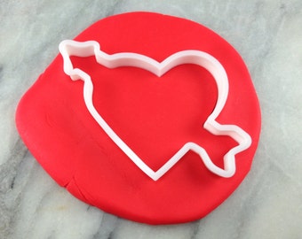 May include: A white plastic cookie cutter in the shape of a heart with an arrow through it, on top of red fondant.