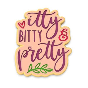 May include: A pink and yellow cookie with the text "itty bitty & pretty" in purple script. A small red heart and a green leaf design are also included.