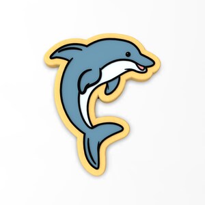 May include: A yellow outlined cookie shaped like a blue dolphin jumping out of the water. The dolphin has a white belly and a black outline.