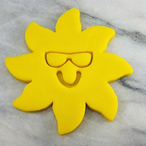 May include: A yellow sun cookie cutter with a smiling face and sunglasses. The cookie cutter is made of plastic and is designed to cut out cookies in the shape of a sun.