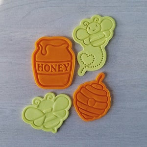 May include: Four cookie cutters in orange and yellow. The cutters are shaped like a honey pot, a bee, a honeycomb, and a bee with a heart-shaped tail.