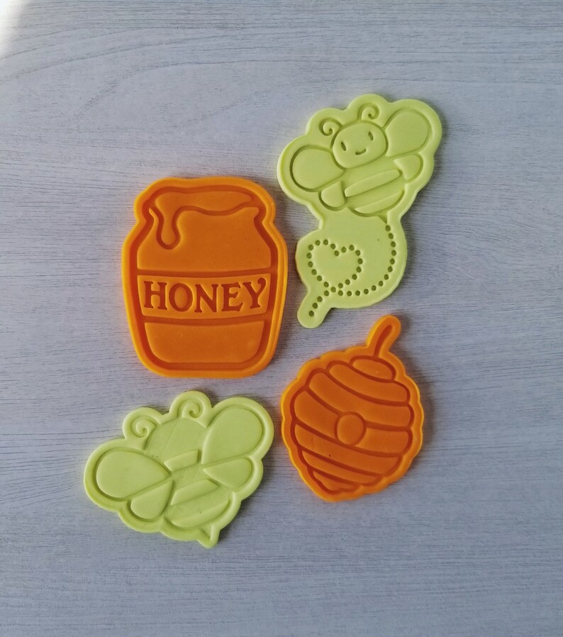 May include: Four cookie cutters in orange and yellow. The cutters are shaped like a honey pot, a bee, a honeycomb, and a bee with a heart-shaped tail.