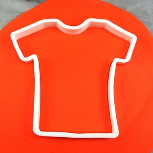 May include: White plastic cookie cutter shaped like a t-shirt on a red surface.