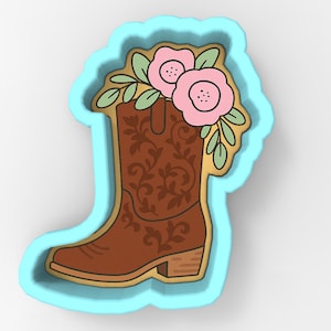 May include: A brown cowboy boot cookie cutter with floral accents. The boot features a detailed floral design and is topped with pink flowers and green leaves. The cutter is outlined in gold and set against a light blue background.