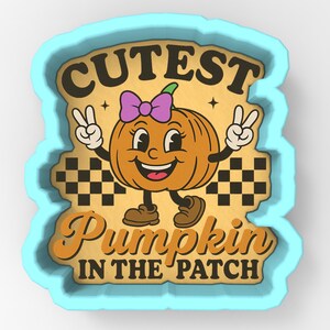 May include: A light blue pumpkin-shaped cookie cutter with the words "CUTEST PUMPKIN IN THE PATCH" and a cartoon pumpkin character with a purple bow, smiling face, and peace signs.